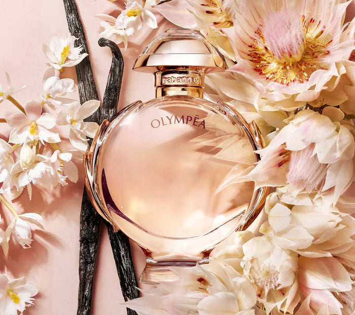 Perfume bottle labeled 'Olympea' surrounded by flowers on a pink background